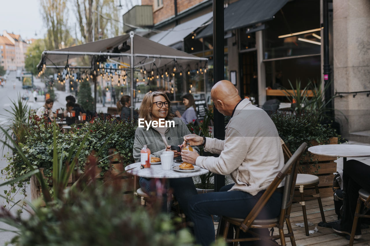 Happy male and female senior friends talking to each other while having food at sidewalk cafe