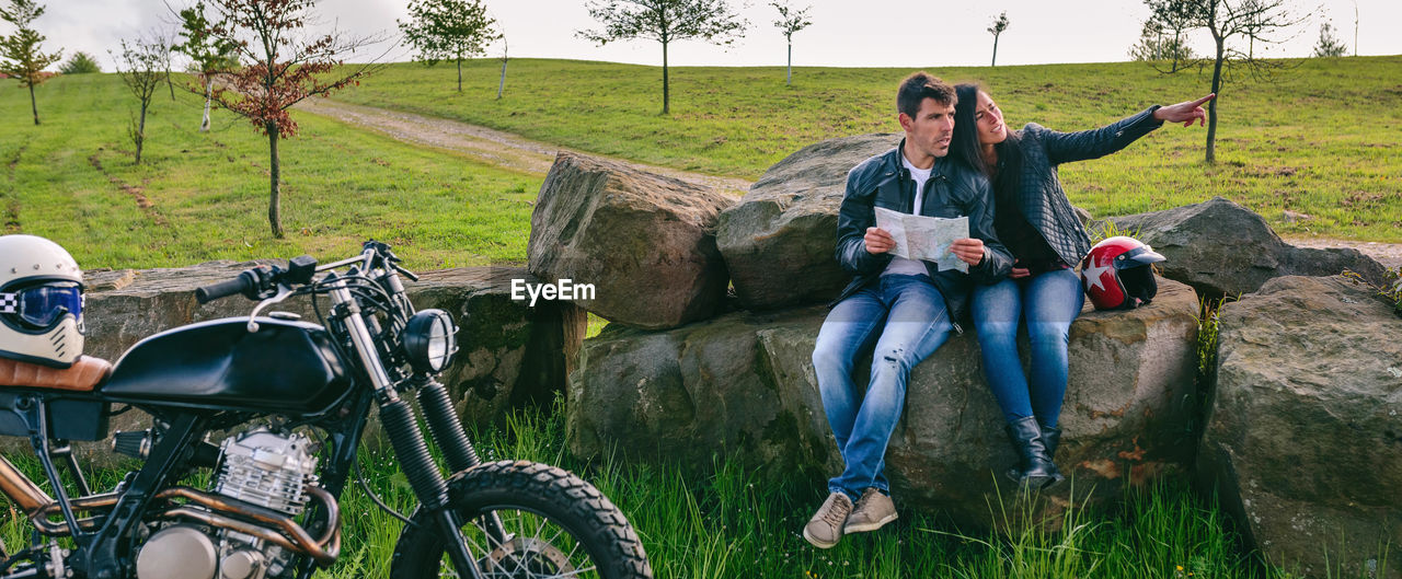 Panoramic shot of bikers sitting on rocks while holding map on land