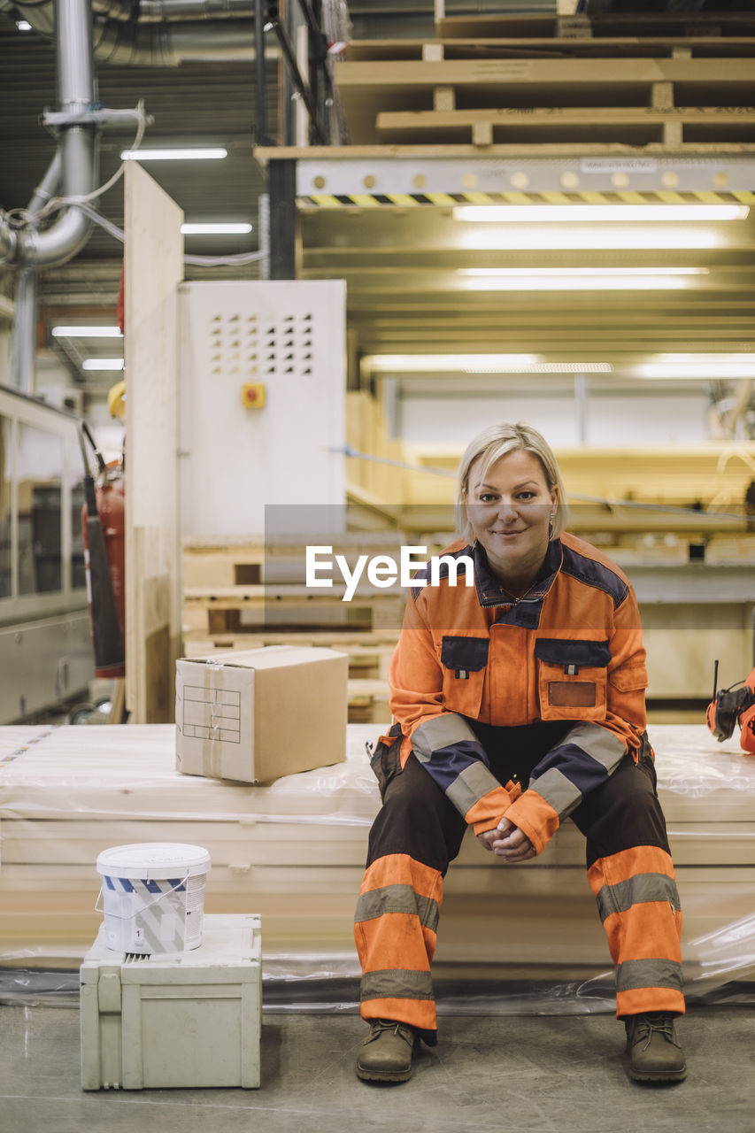 Full length portrait of smiling mature carpenter wearing reflective clothing while sitting in warehouse