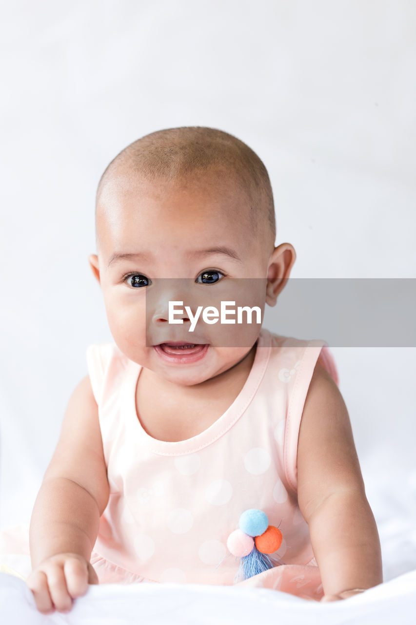 Close-up portrait of cute baby girl sitting on bed against wall at home