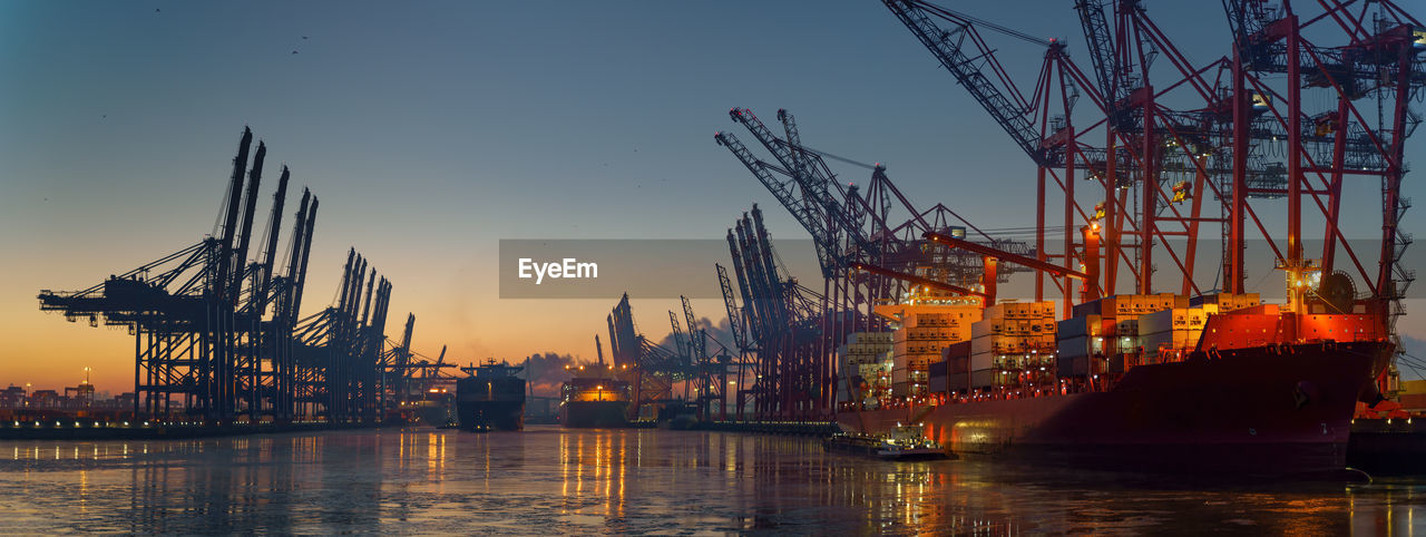 Cranes at commercial dock against sky during sunset