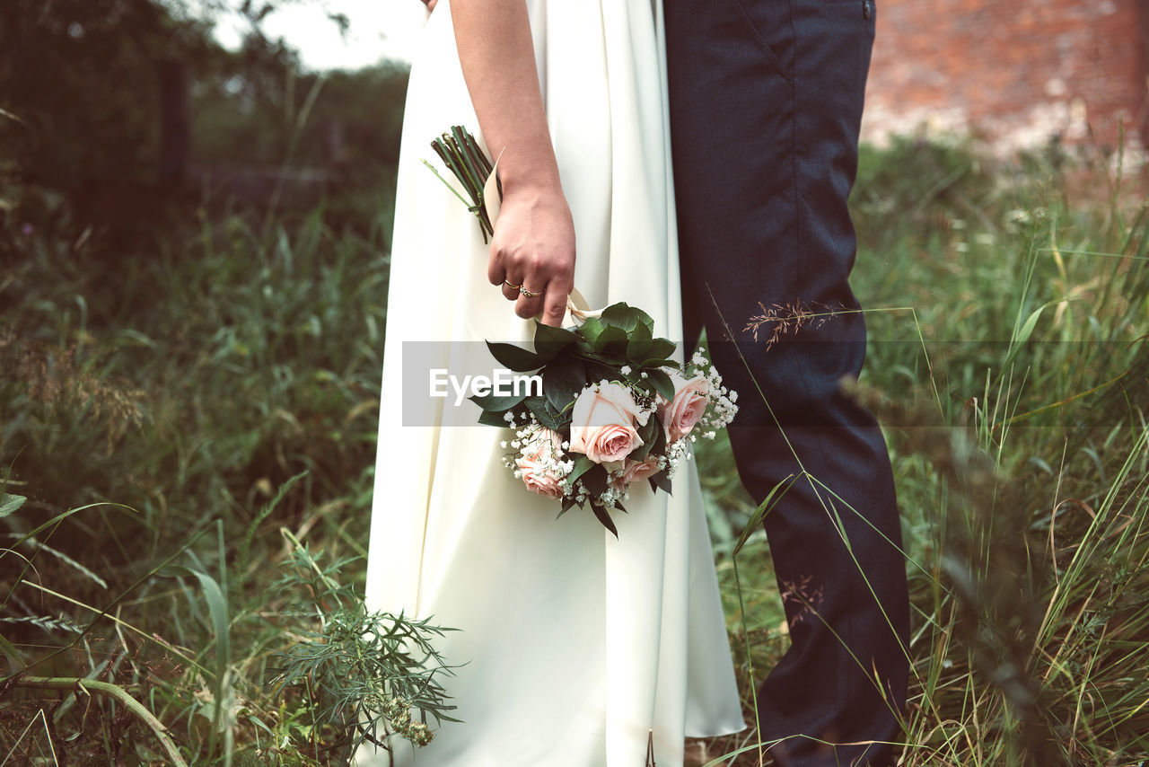 Midsection of bride with flower bouquet standing by groom over field