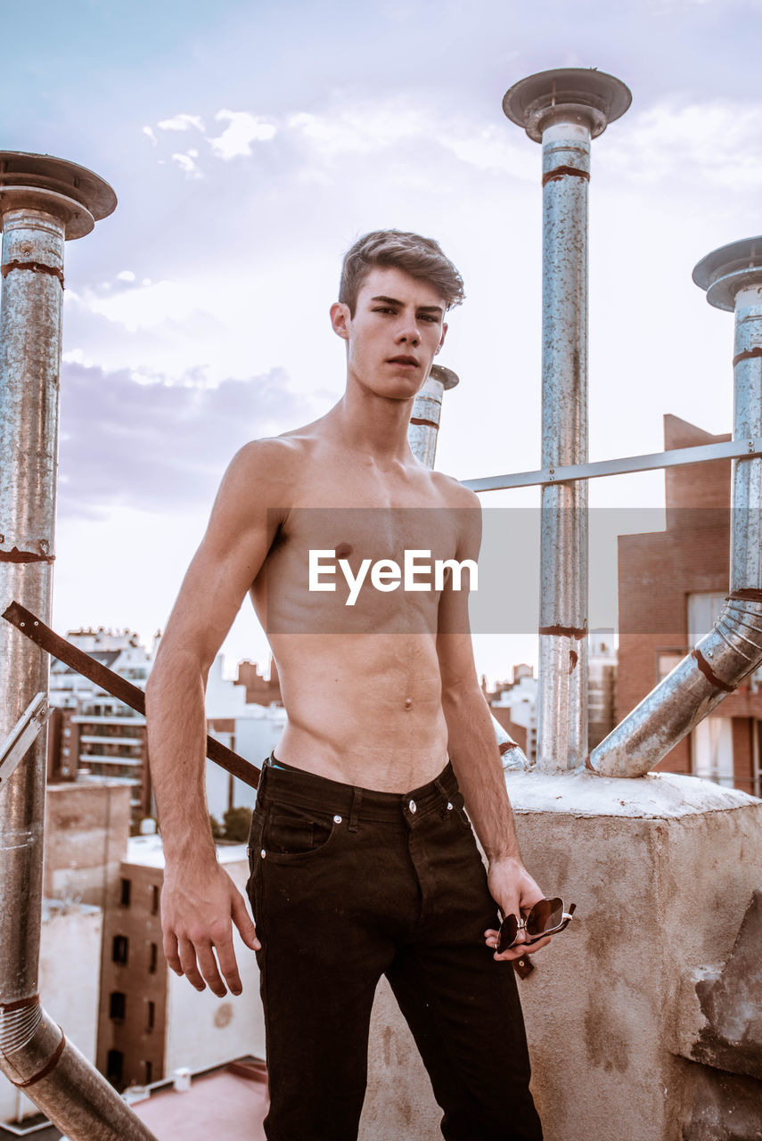 Portrait of shirtless young man standing against buildings