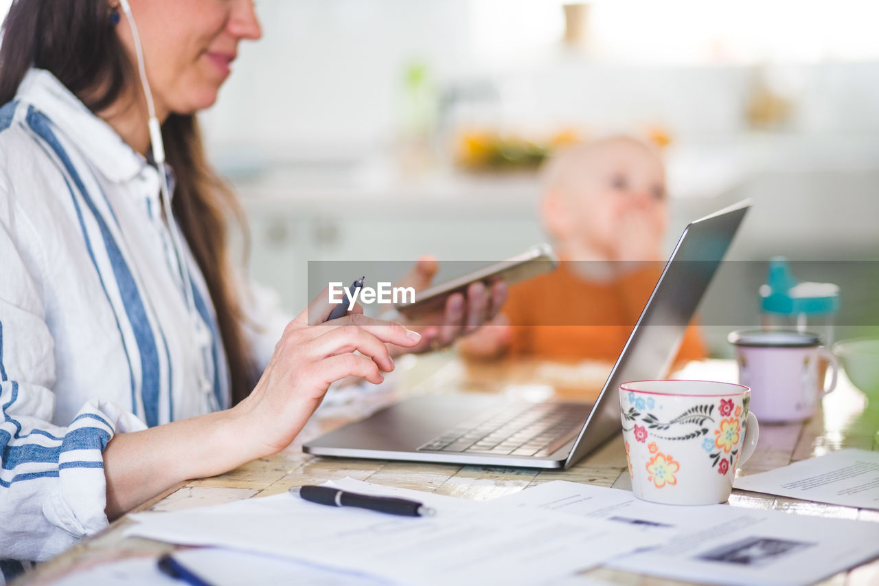 Midsection of working mother using technologies while daughter sitting in background at dining table