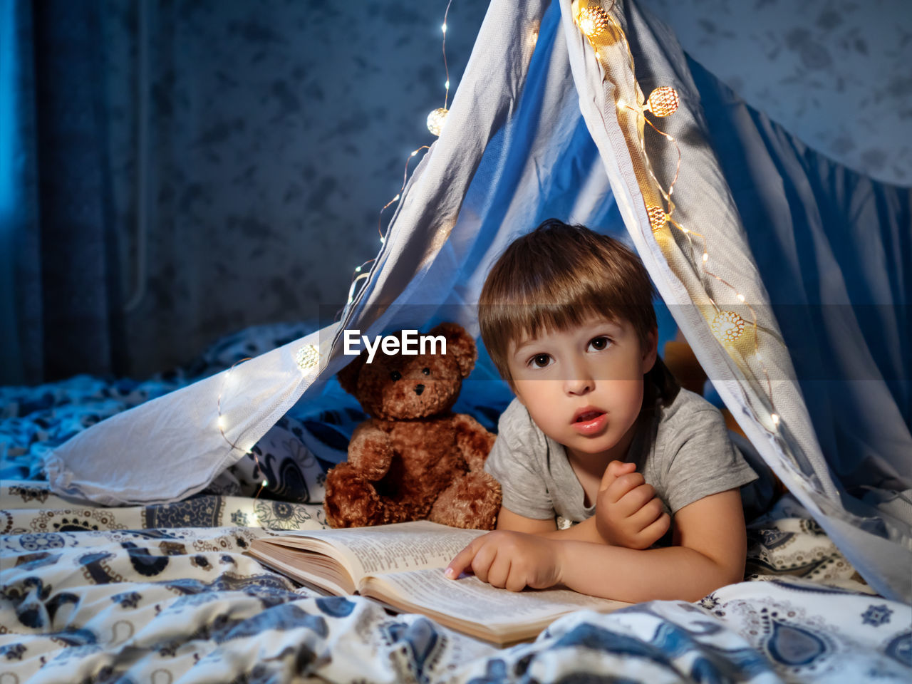 Little boy reads book. toddler plays in tent made of linen sheet on bed. cozy evening with  book.