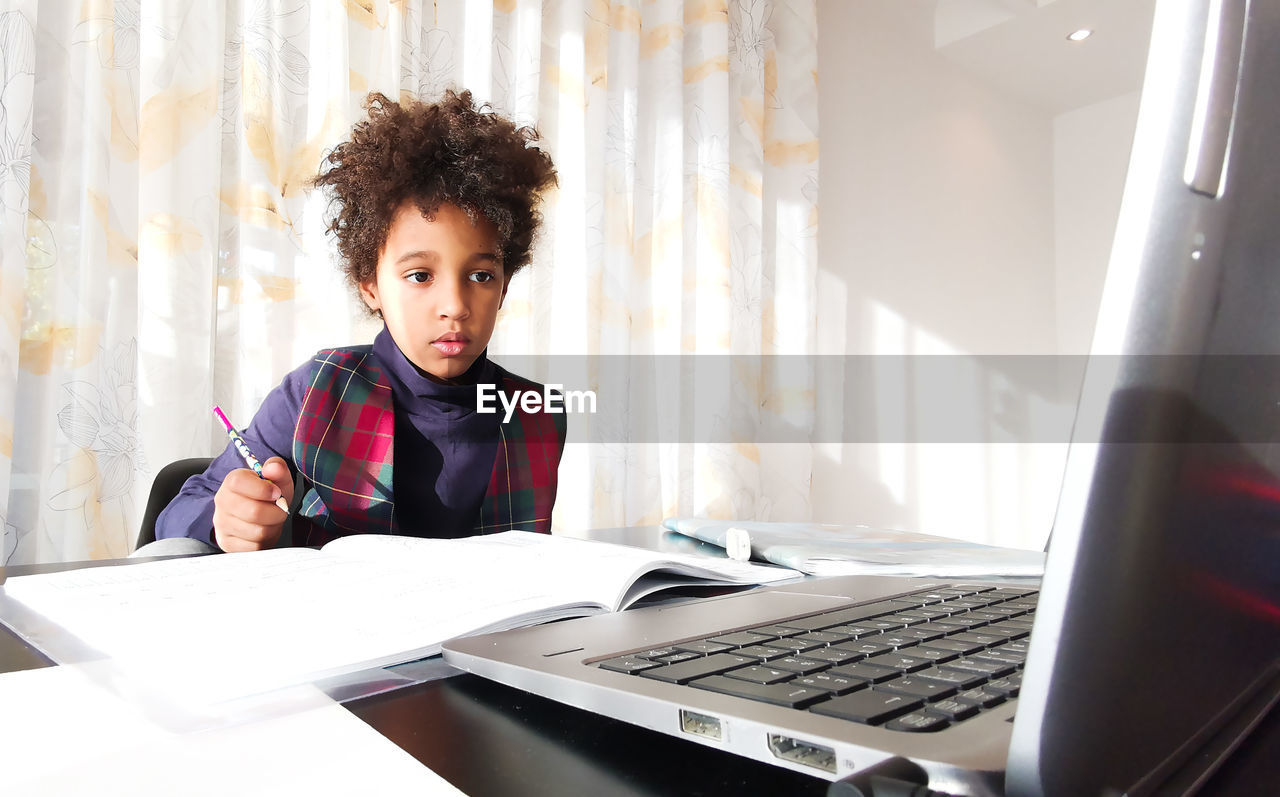 Mixed race kid with curly hair doing remote homework in front of laptop