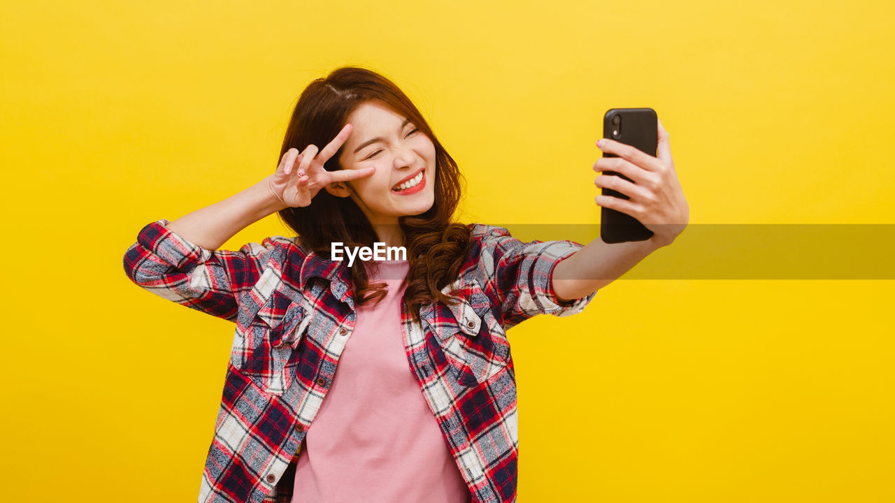 Smiling young woman talking selfie with mobile phone against yellow background