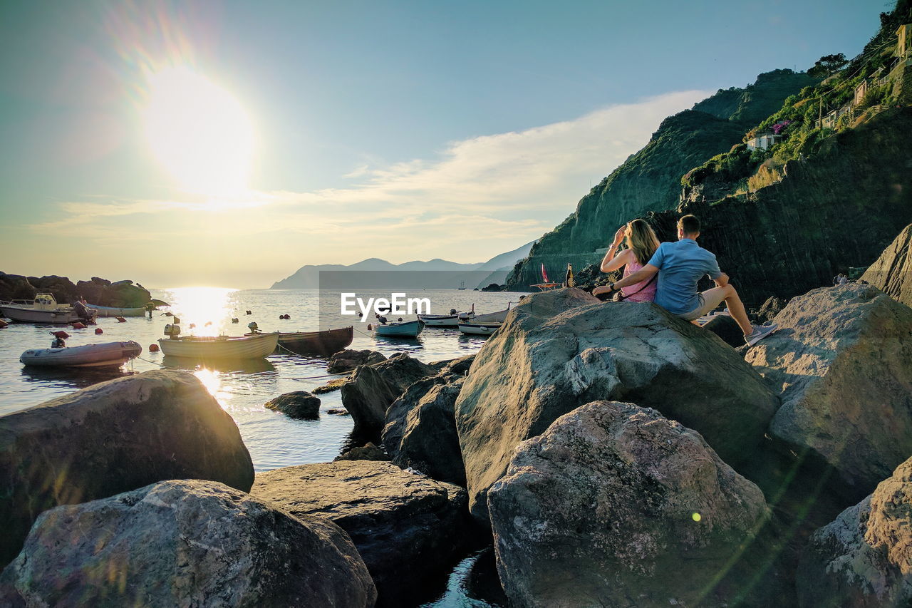 PEOPLE SITTING ON ROCK BY SEA AGAINST SKY