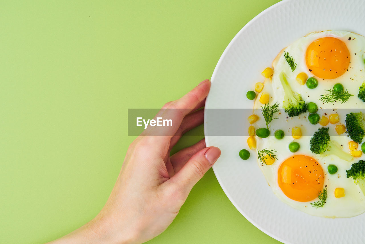 Female hand touching plate with fried eggs, broccoli, corn on light green background