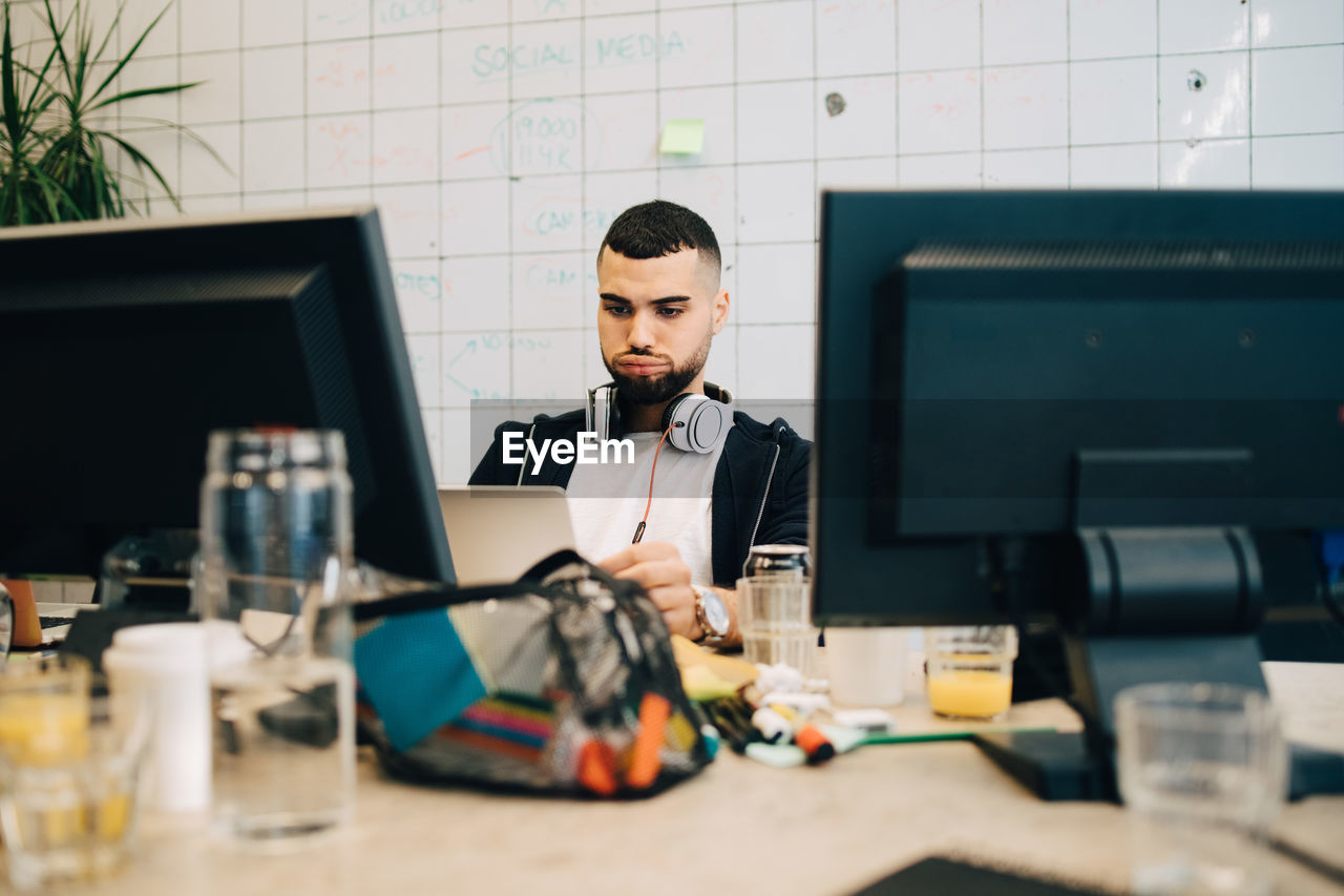 Young male computer hacker making face while coding on laptop at creative office