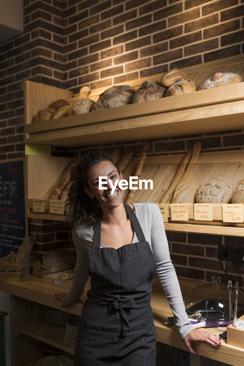 Smiling young owner standing against baked food rack in illuminated bakery