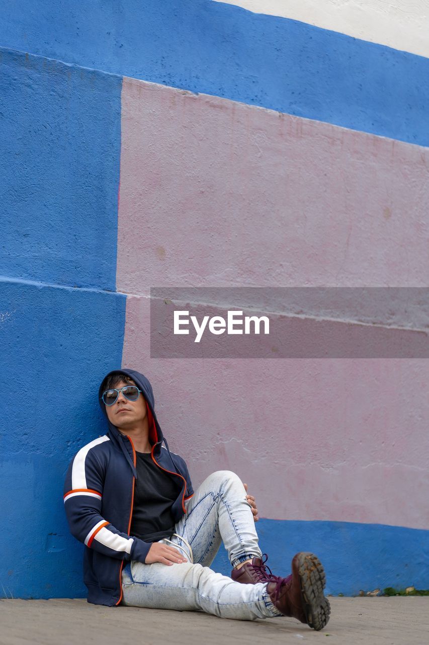 Man sitted on the floor wearing a  hoodie and sunglasses with a colorful wall as background.