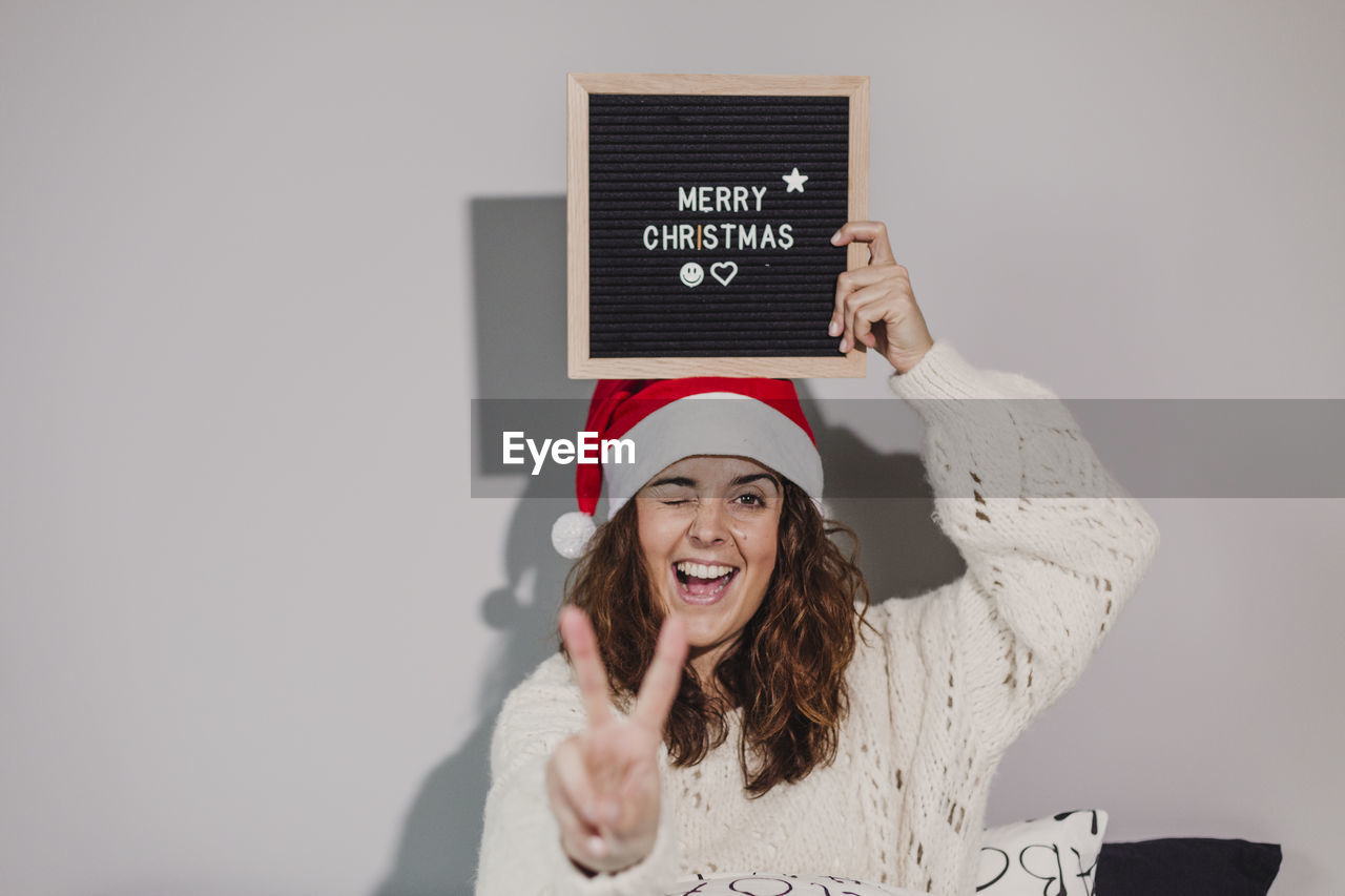 Portrait of woman winking and gesturing with merry christmas text at home