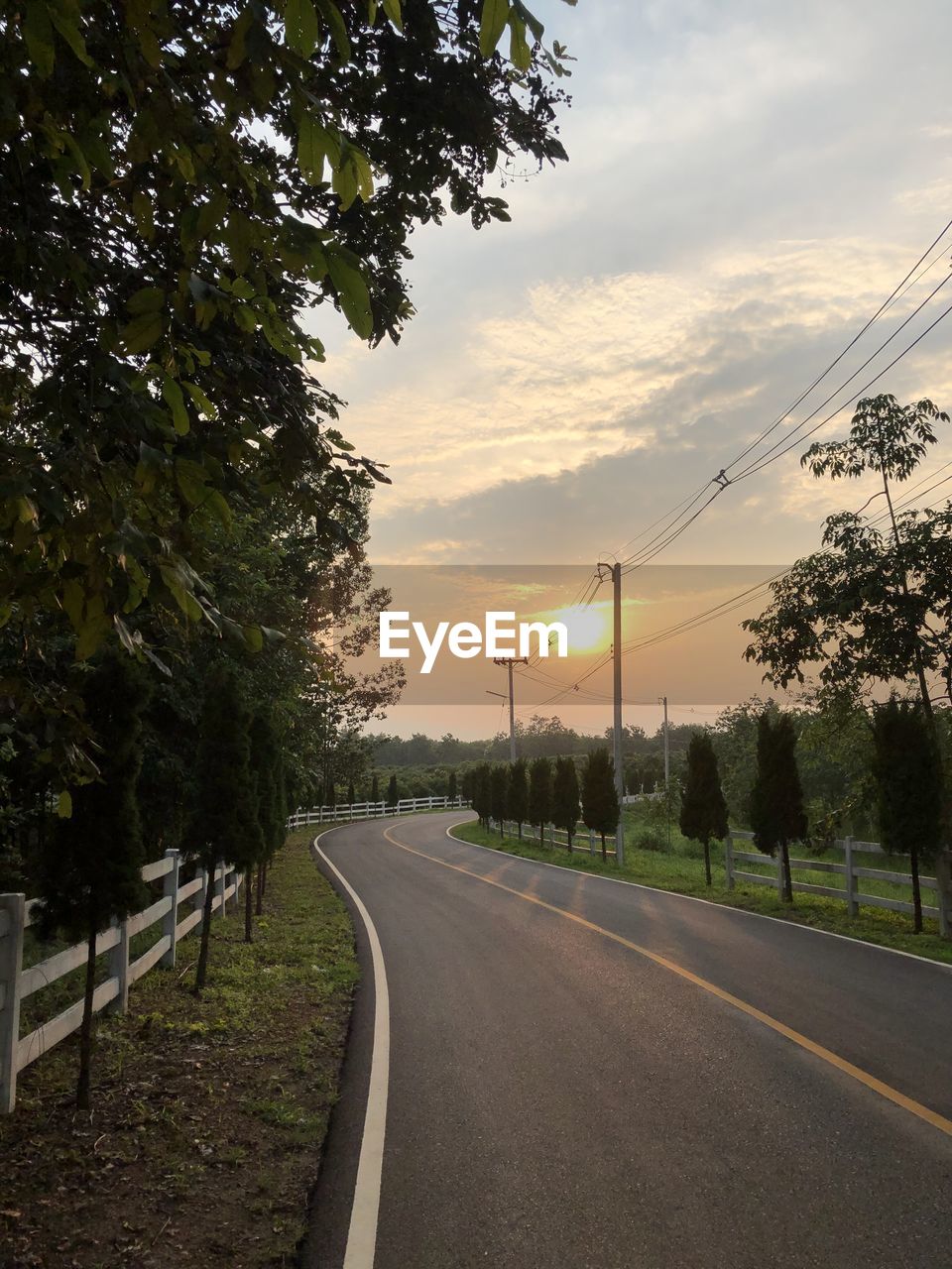 Empty road by trees against sky during sunset