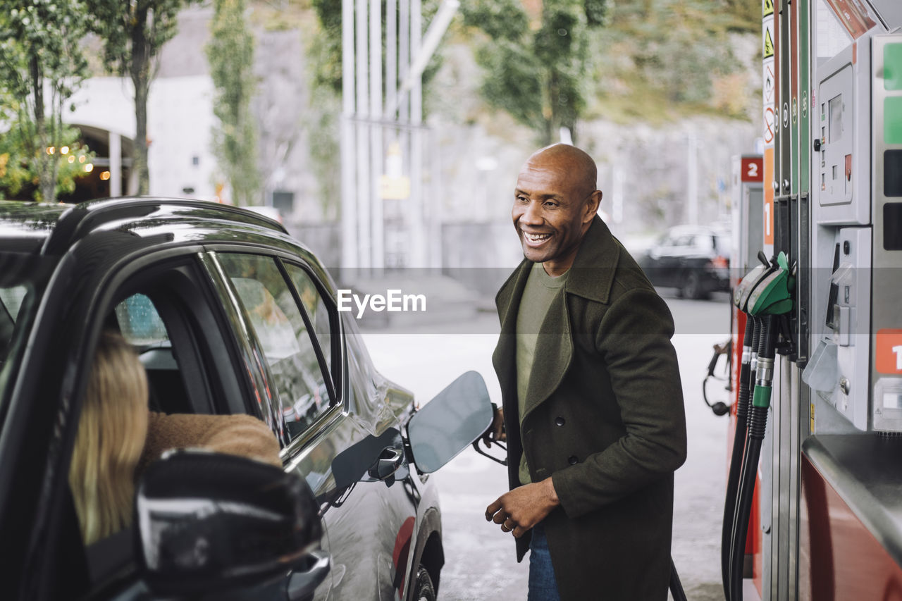 Man with shaved head talking to woman while refueling car while at gas station