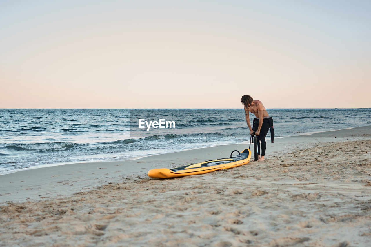 Man in wetsuit inflating paddle board at the seashore on sandy beach