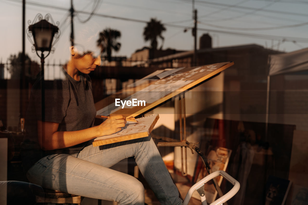 Side view through the glass of a woman drawing at home at sunset