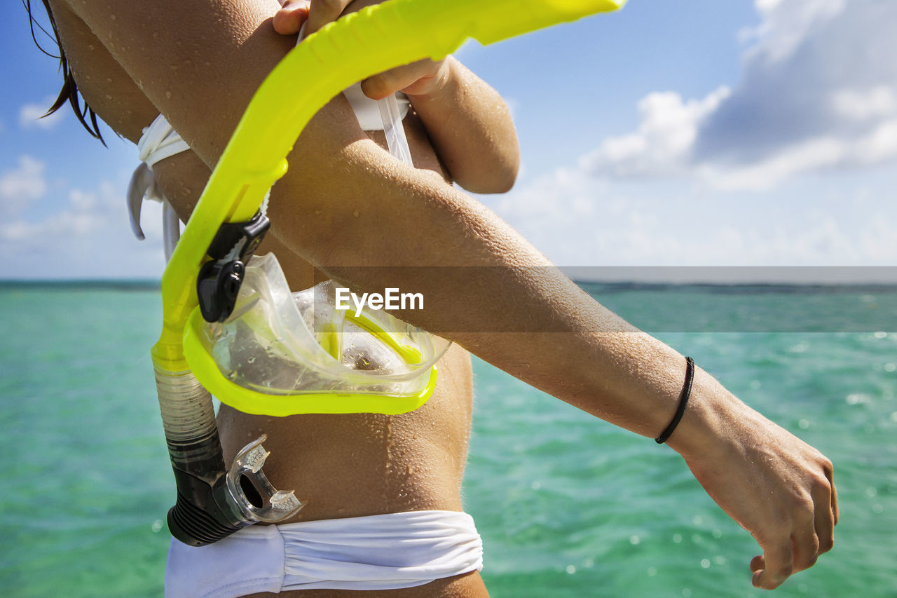 Midsection of woman holding snorkel while standing in sea against cloudy sky
