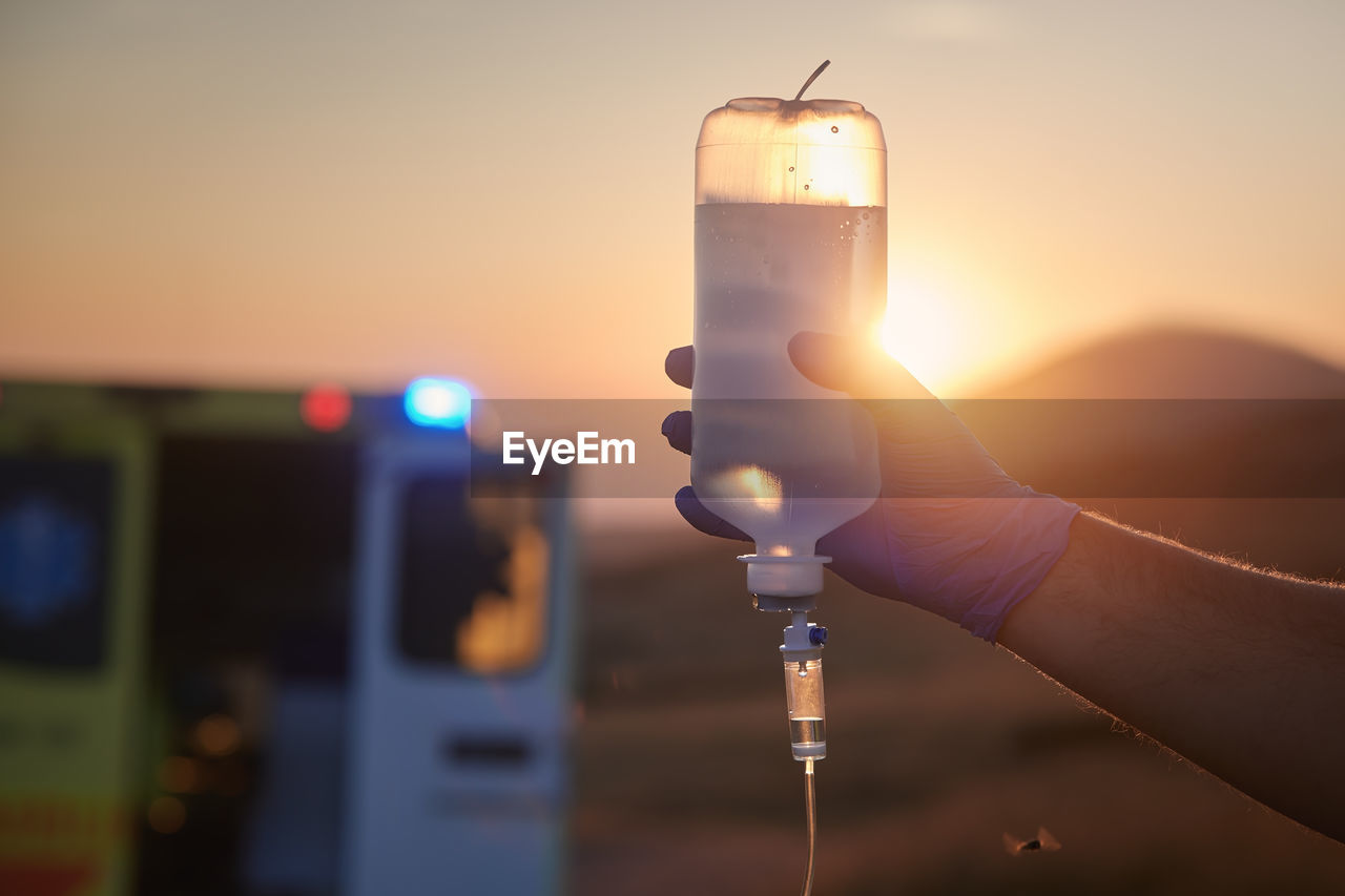 Close-up hand of paramedic holding infusion set against ambulance car of emergency medical service.