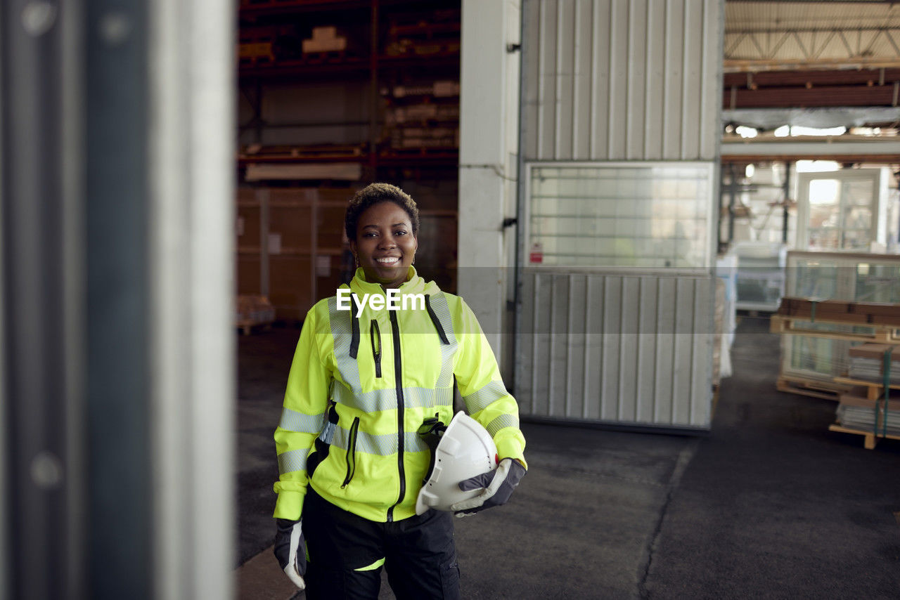 Portrait of smiling blue-collar worker in protective workwear holding hardhat in factory