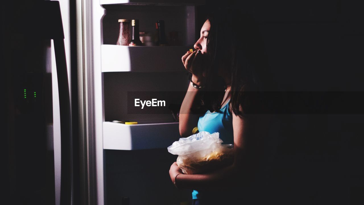 Side view of teenage girl eating while standing in dark at home