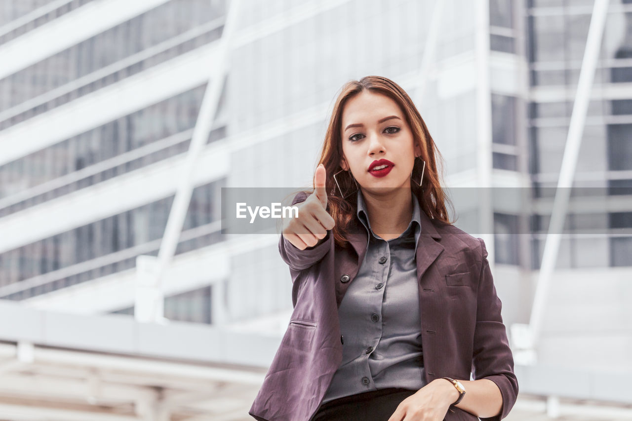 Portrait of young businesswoman showing thumbs up against buildings in city