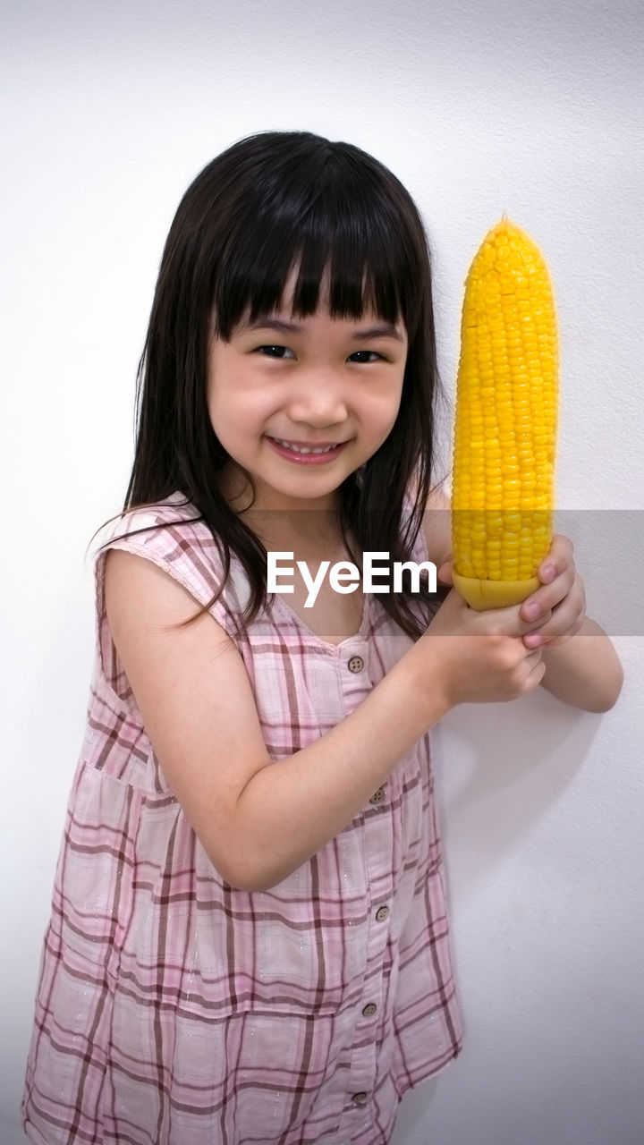 Portrait of smiling girl holding corn while standing against wall