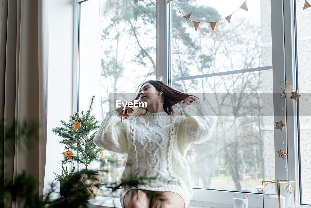 Woman in warm white winter sweater sitting on the window at home at christmas eve holding cup