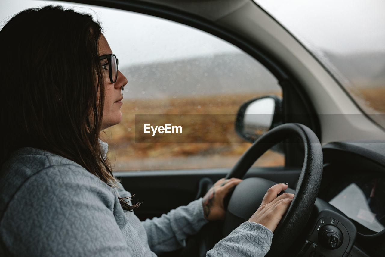 Side view of young woman in eyeglasses with piercing driving automobile between wild lands with hills in rainy weather