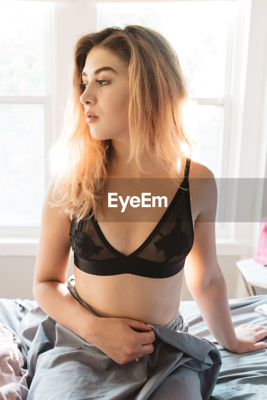 Thoughtful woman wearing black bra sitting on bed during sunny day