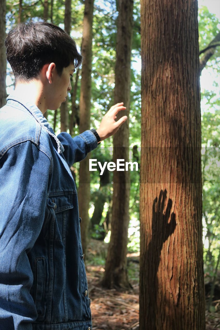 Side view of young man touching tree in forest