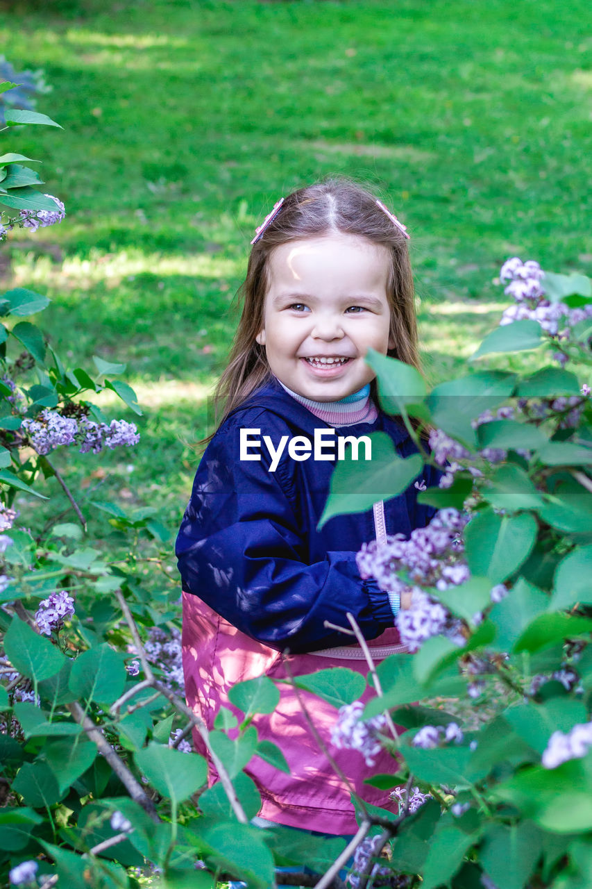 child, childhood, smiling, plant, one person, happiness, flower, nature, women, female, portrait, emotion, looking at camera, flowering plant, leisure activity, day, growth, green, blond hair, teeth, smile, cute, innocence, cheerful, front view, outdoors, casual clothing, freshness, three quarter length, long hair, toddler, field, hairstyle, lifestyles, clothing, enjoyment, meadow, garden, grass, spring, standing, leaf, land, positive emotion, person, plant part, fun, beauty in nature