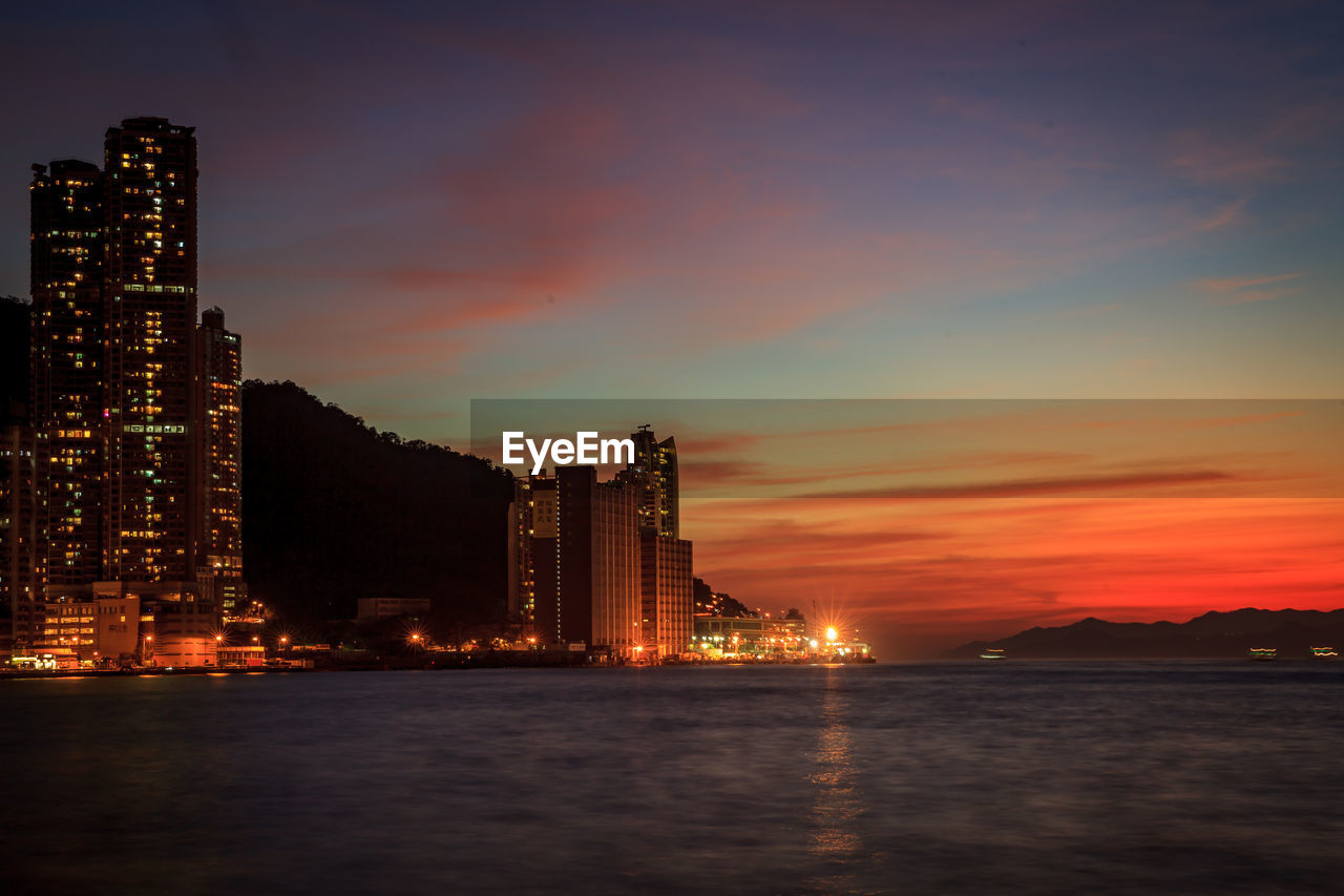 SEA BY ILLUMINATED BUILDINGS AGAINST SKY AT SUNSET