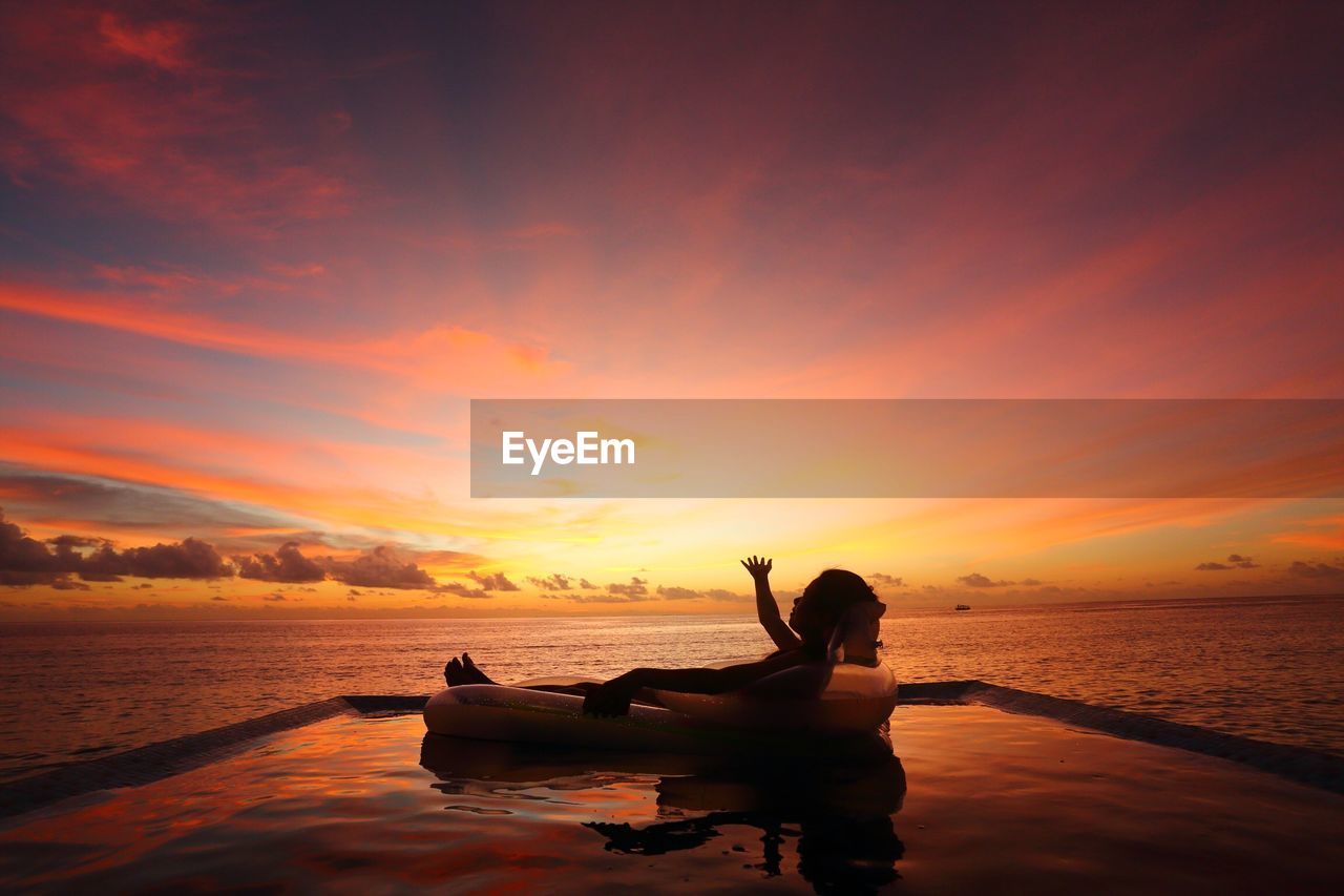 Side view of woman floating on inflatable raft by sea against dramatic sky during sunset