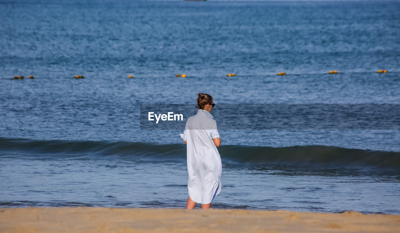 REAR VIEW OF WOMAN STANDING ON BEACH AGAINST SKY