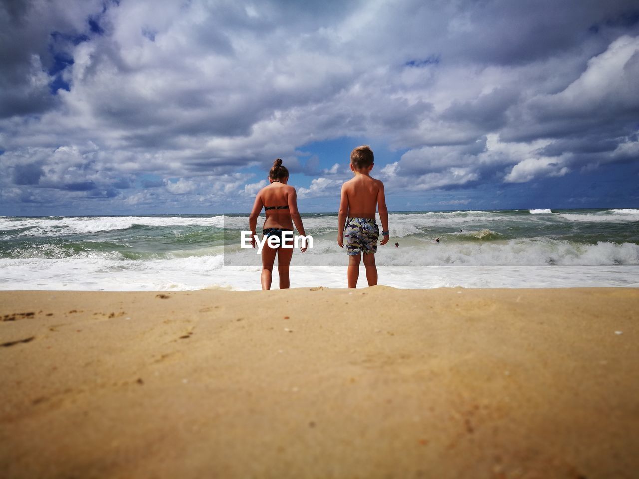Rear view of siblings standing on beach against cloudy sky