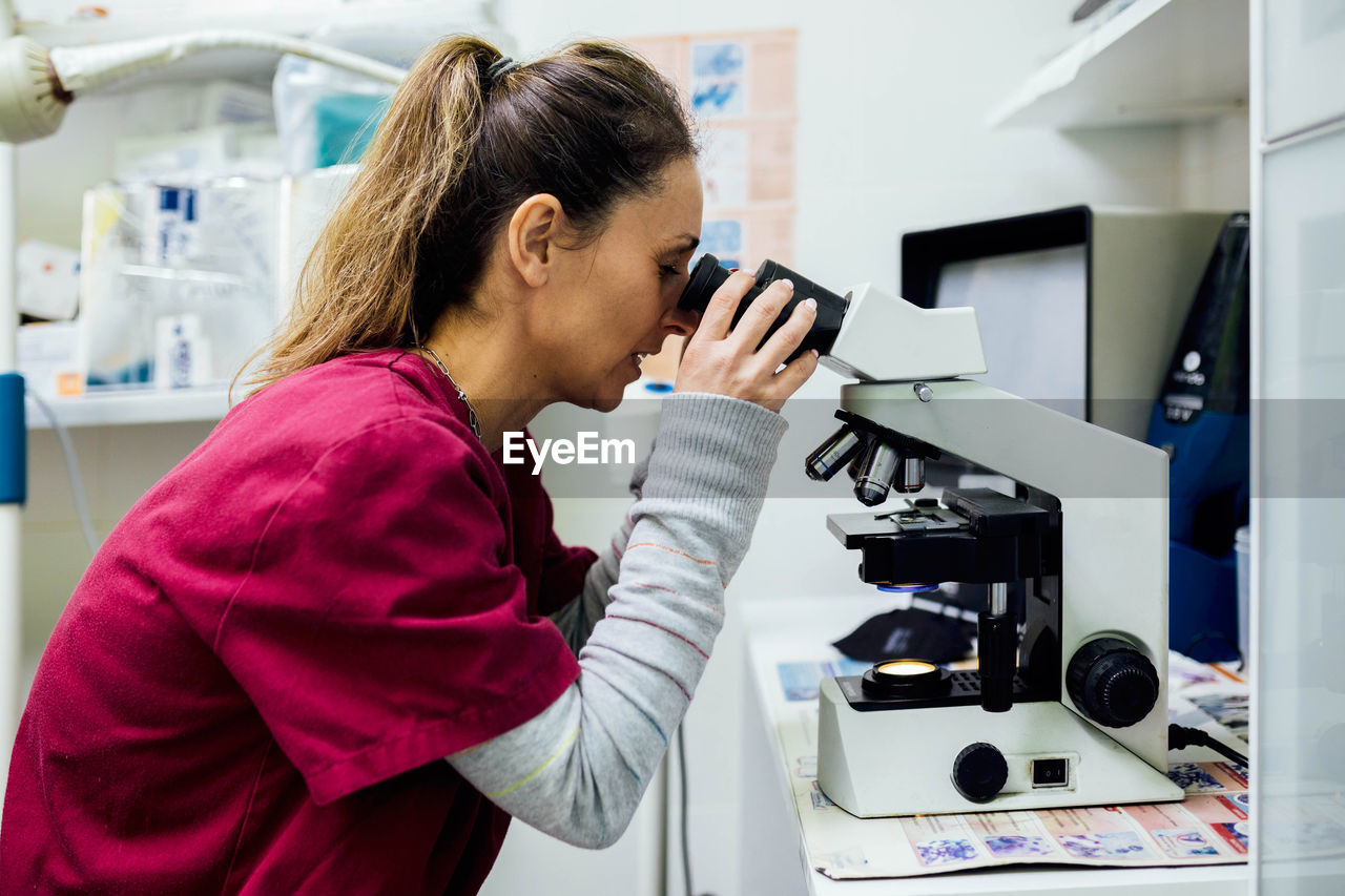 Side view of focused female veterinarian in uniform looking through microscope while checking results of medical procedure in modern clinic