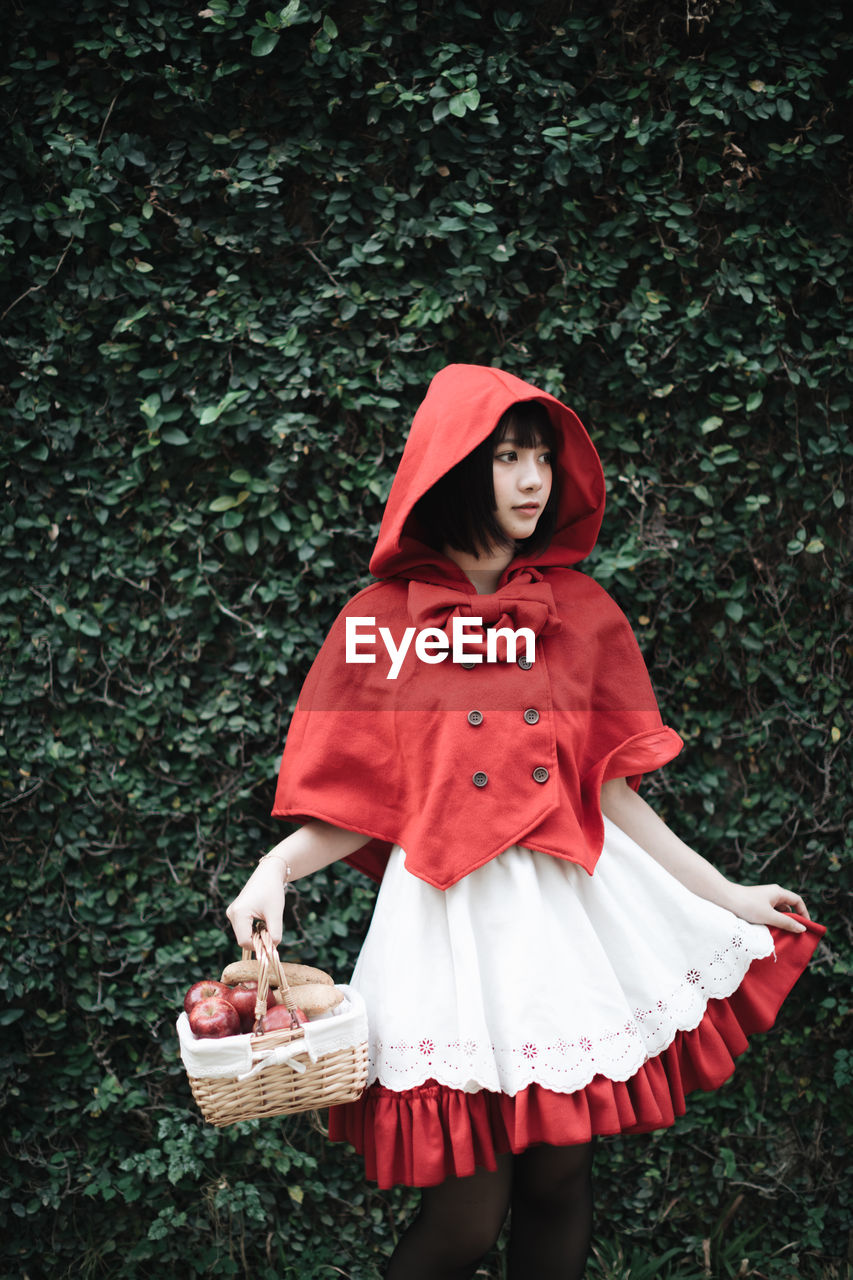 Woman in little red riding hood costume holding basket while standing against plants