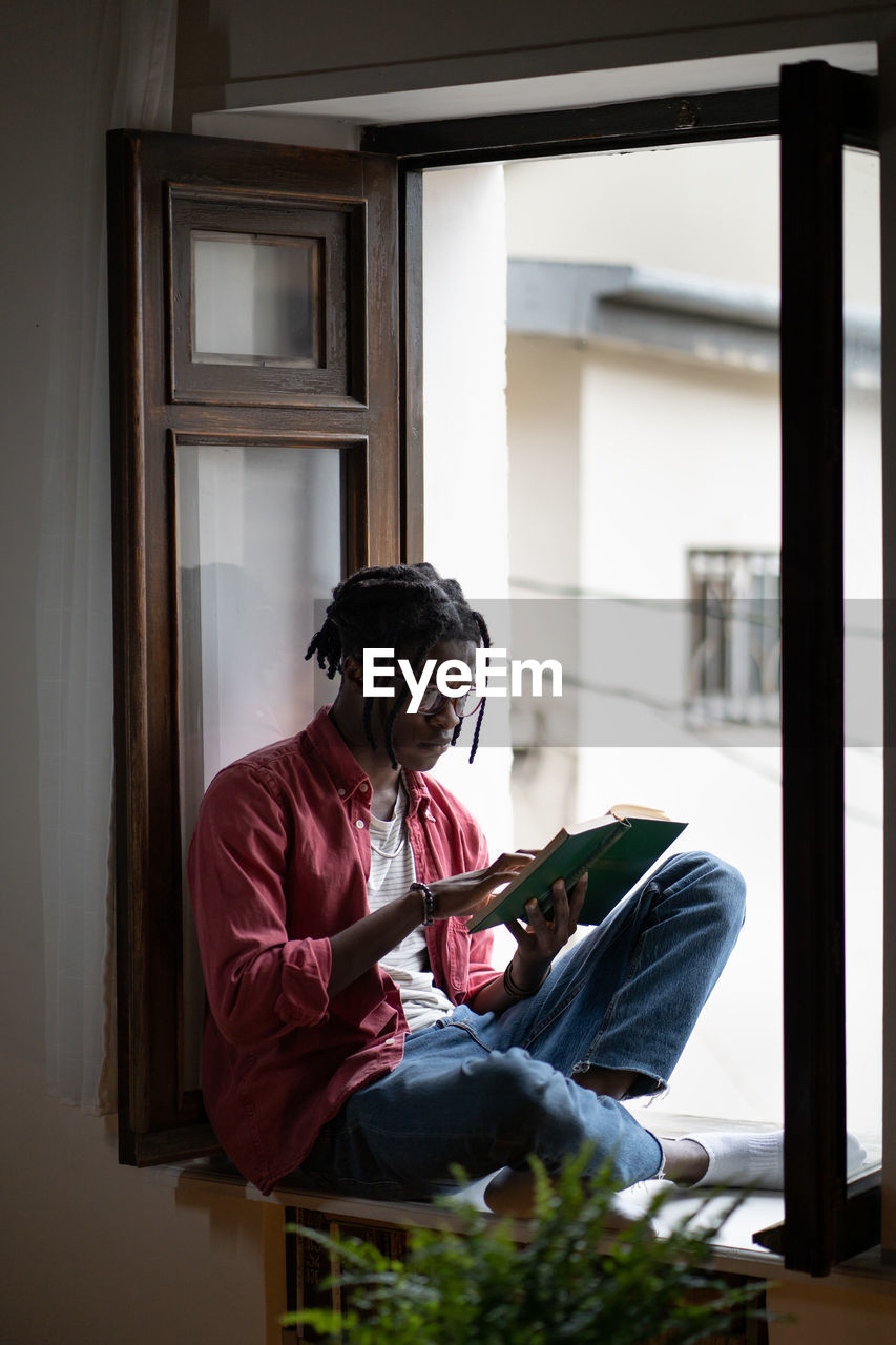 Upset black man excited from reading sits on windowsill with dramatic book worries about characters.