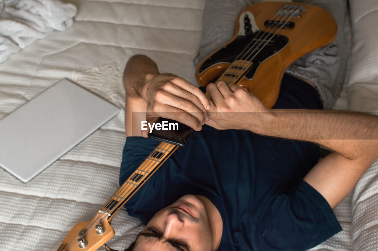 Man lying on a bed using a mobile phone after playing electric bass.