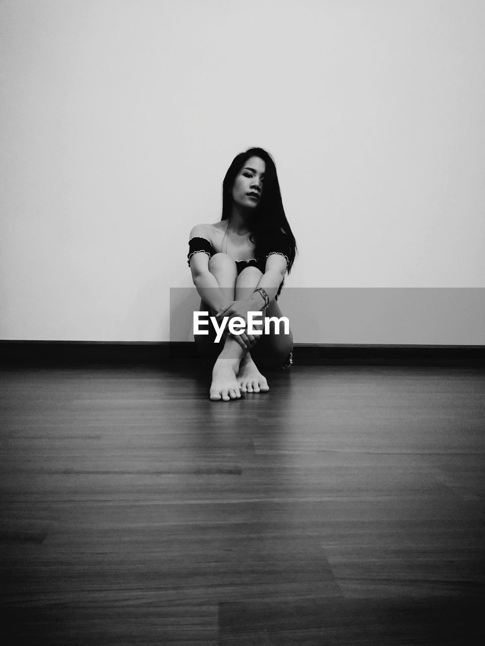 Full length portrait of young woman sitting on hardwood floor against white wall at home