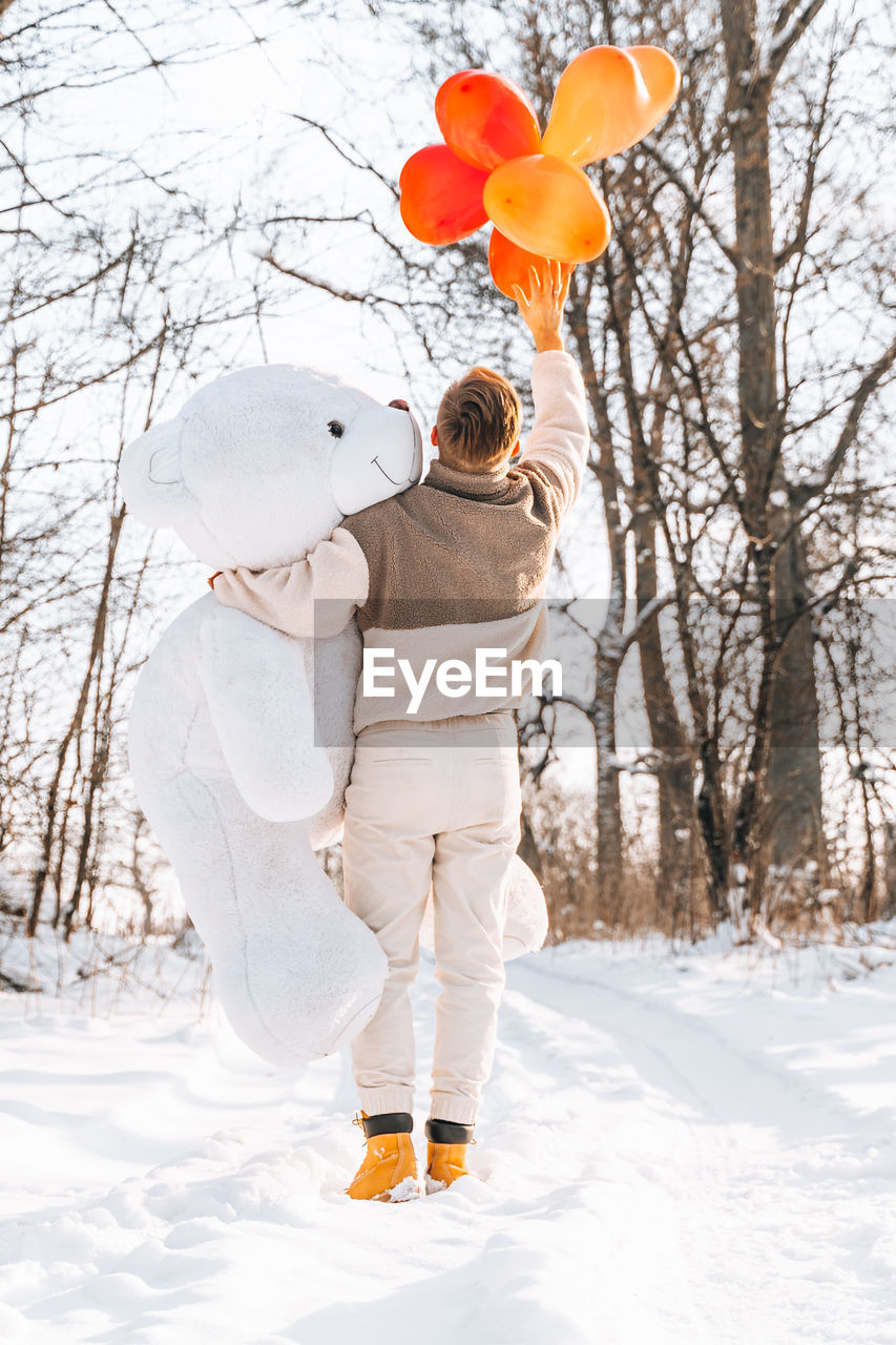 Rear view of man holding stuffed toy with balloon standing on snow covered land