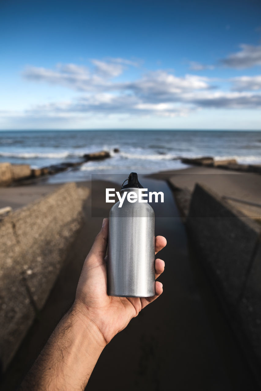 Cropped hand of person holding bottle against sea and sky