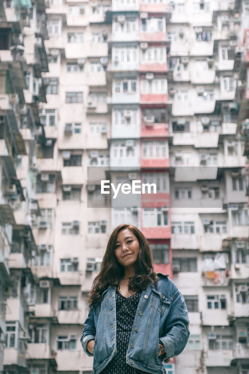 PORTRAIT OF SMILING YOUNG WOMAN STANDING AGAINST CITY