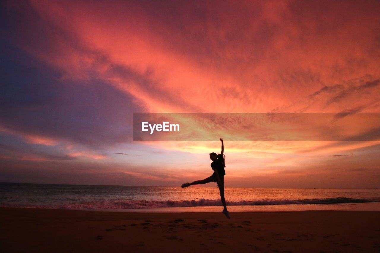 Silhouette person jumping on beach against multi colored cloudy sky