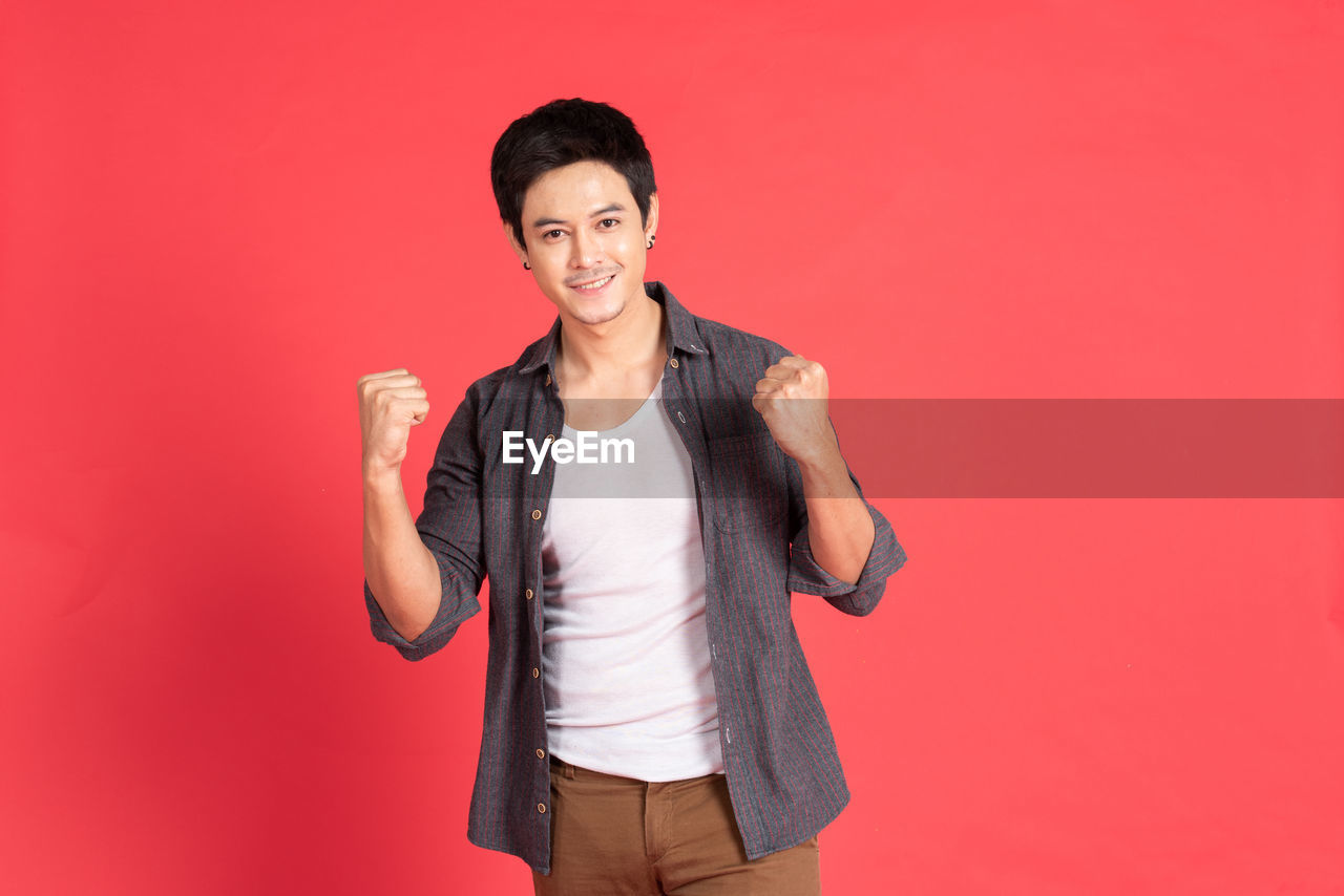 Happy young asian man in casual dress celebrates with two hand on isolate red background. 