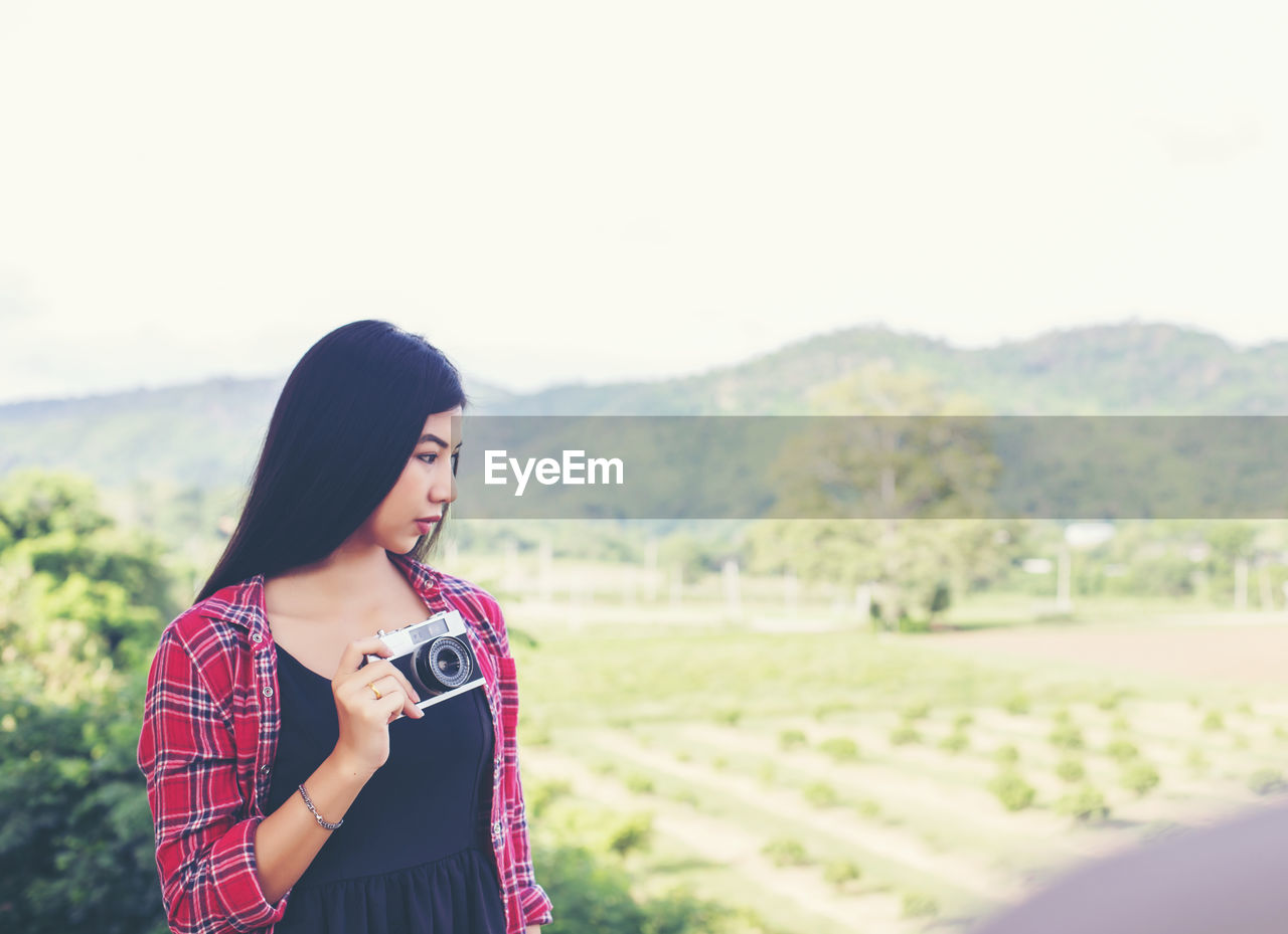 Young woman holding camera while standing on land against clear sky