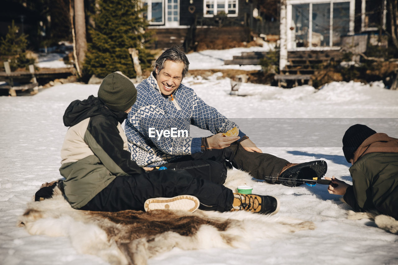 Smiling father talking with son while boy fishing lying on snow during sunny day