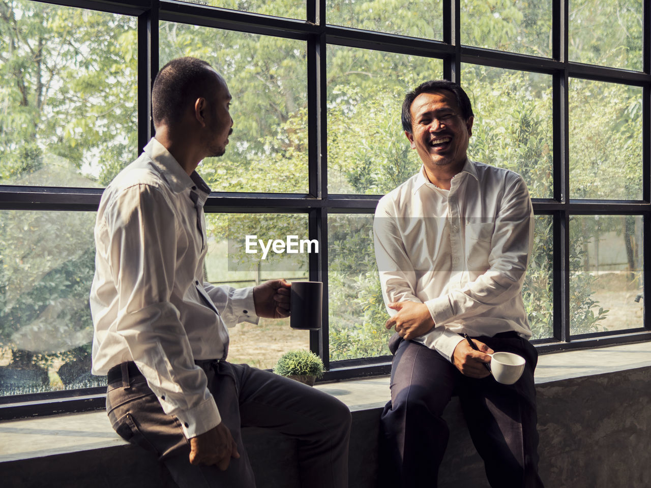 Cheerful businessman taking coffee break with colleague by window in office