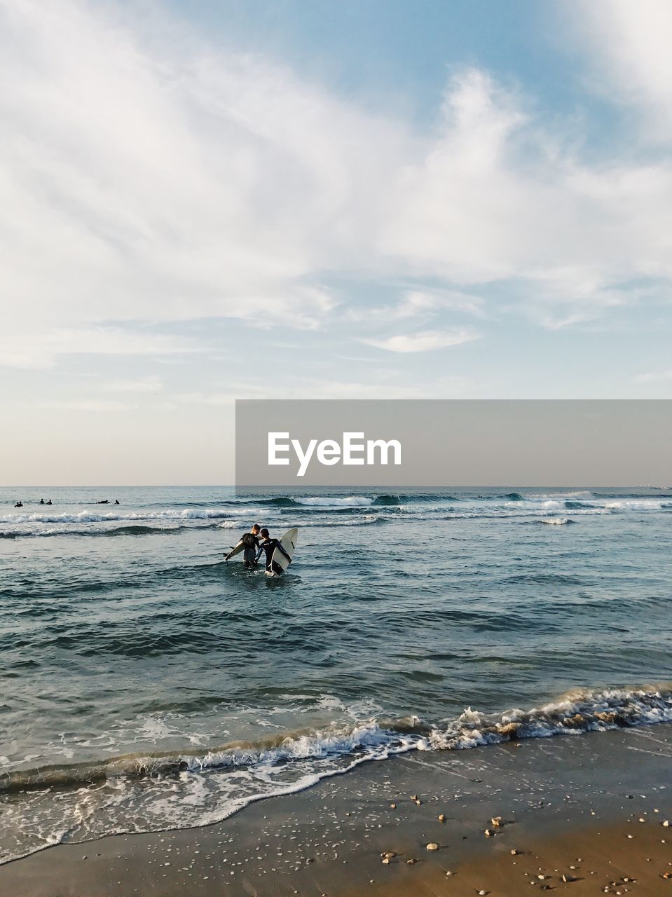 Friends with surfboards walking in sea against cloudy sky